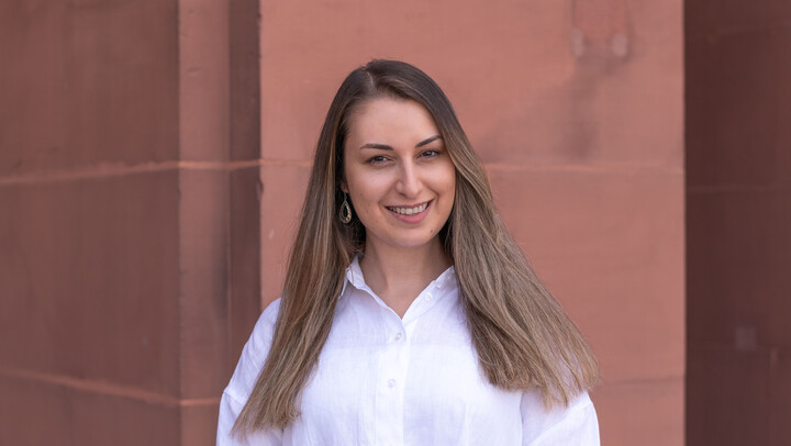 Samia Hanna. She is wearing a white shirt and is standing in front of a brown stone wall.