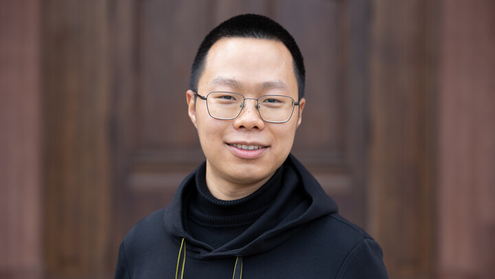 Siyu Zhang. He is wearing a black jacket and is standing in front of a brown door.