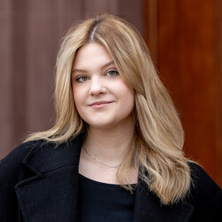 Porträt von Emilia Toth. Emilia Toth trägt einen schwarzen Mantel und ein schwarzes Oberteil. Sie hat blonde Haare, die sie offen trägt und die ihr über die Schultern reichen. Um den Hals trägt sie eine dezente silberne Kette. Sie schaut offen in die Kamera.ie Kamera.