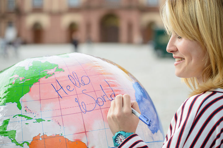 Lächelnde Frau in einem gestreiften Shirt auf dem Ehrenhof, schreibt mit einem Stift auf einen großen Globus "Hello World".