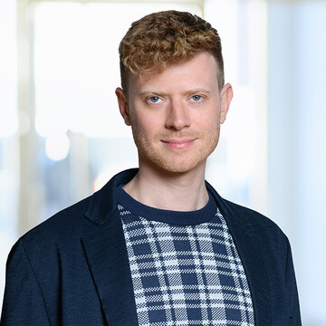 Moritz Klenk hat kurze lockige blonde Haare. Er trägt ein blau-weiß-kariertes T-Shirt und ein blaues Jacket.