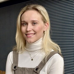 Porträt von Anna-Lena Baumann. Anna-Lena Baumann trägt eine braun-karrierte Latzhose, einen hellen Rolli und dezenten Goldschmuck. Sie hat blonde schulterlange Haare, die sie offen trägt. Sie lacht keck in die Kamera.