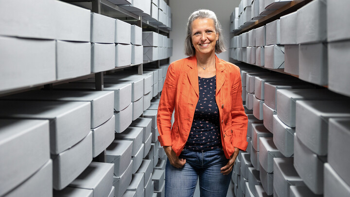 Sandra Eichfelder trägt eine orangene Jacke und steht inmitten von Regalen mit grauen Boxen.