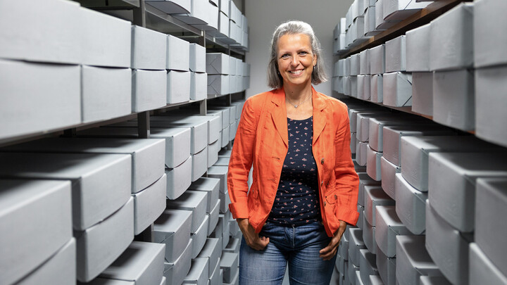 Sandra Eichfelder trägt eine orangene Jacke und steht inmitten von Regalen mit grauen Boxen.