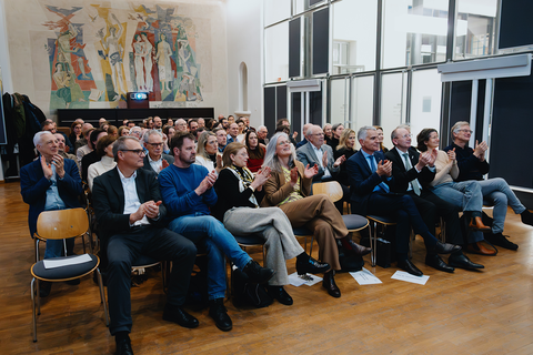 Mehrere Dutzend Personen sitzen in mehreren Reihen auf Stühlen.