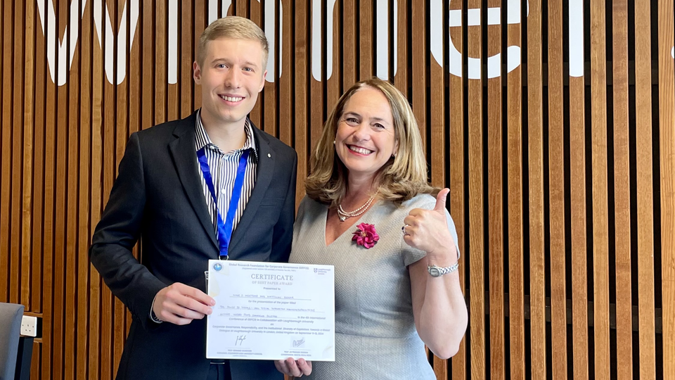 Marc wears a dark suit, is holding an award certificate and is standing next to Professor Ruth V. Aguilera who is wearing a light grey dress.