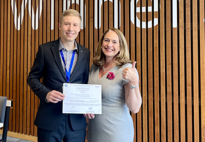 Marc wears a dark suit, is holding an award certificate and is standing next to Professor Ruth V. Aguilera who is wearing a light grey dress.