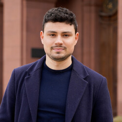 Portrait of Nils Roßnagel. He is wearing a dark blue coat with a matching sweater and has short dark brown hair.