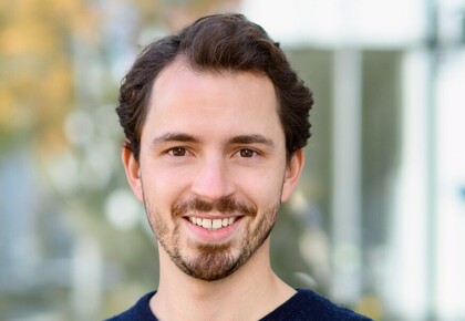 Philipp Kadel hat wellige dunkelbraune Haare und einen Bart. Er steht im Freien vor einem Gebäude und einem Baum und trägt einen dunkelblauen Pullover.