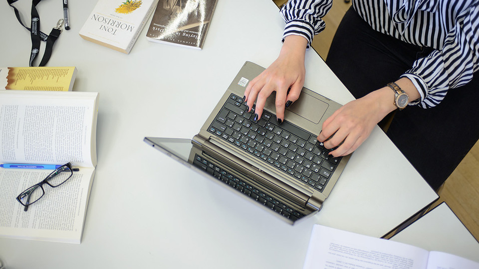 Eine Frau tippt in ihren Laptop und daneben liegen fünf Bücher.