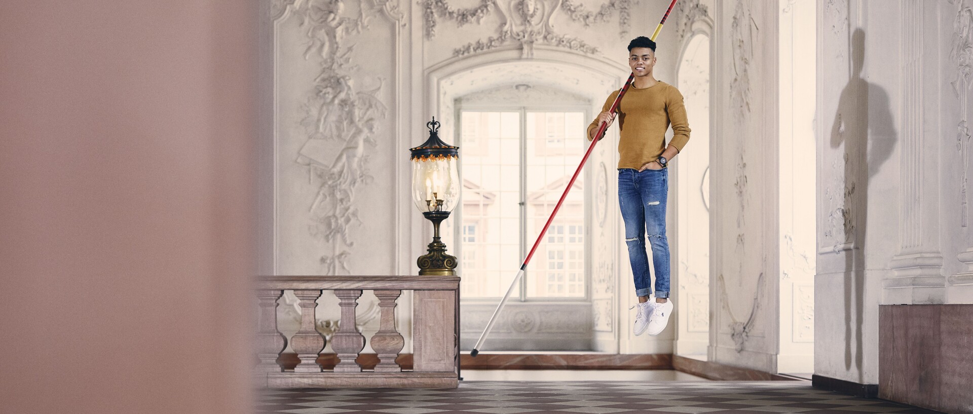 Ein Mann mit einem langen Stab über der Schulter schwebt über dem Boden. Im Hintergrund ein Treppengeländer, große Fenster und ornamierte Wände.