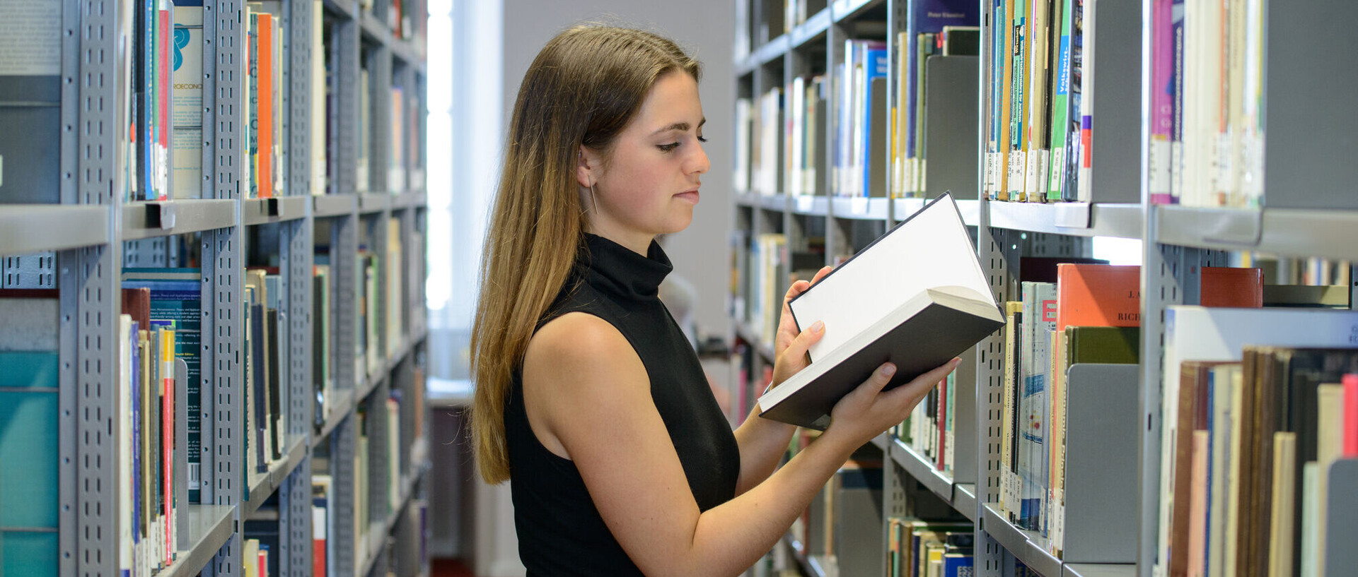 Eine Studentin steht zwischen zwei Bücherregalen und liest in einem Buch.