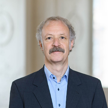 Prof. Dr. Moritz Fleischmann hat graue Haare und einen Schnauzer. Er trägt ein dunkles Jackett und ein gestreiftes Hemd.