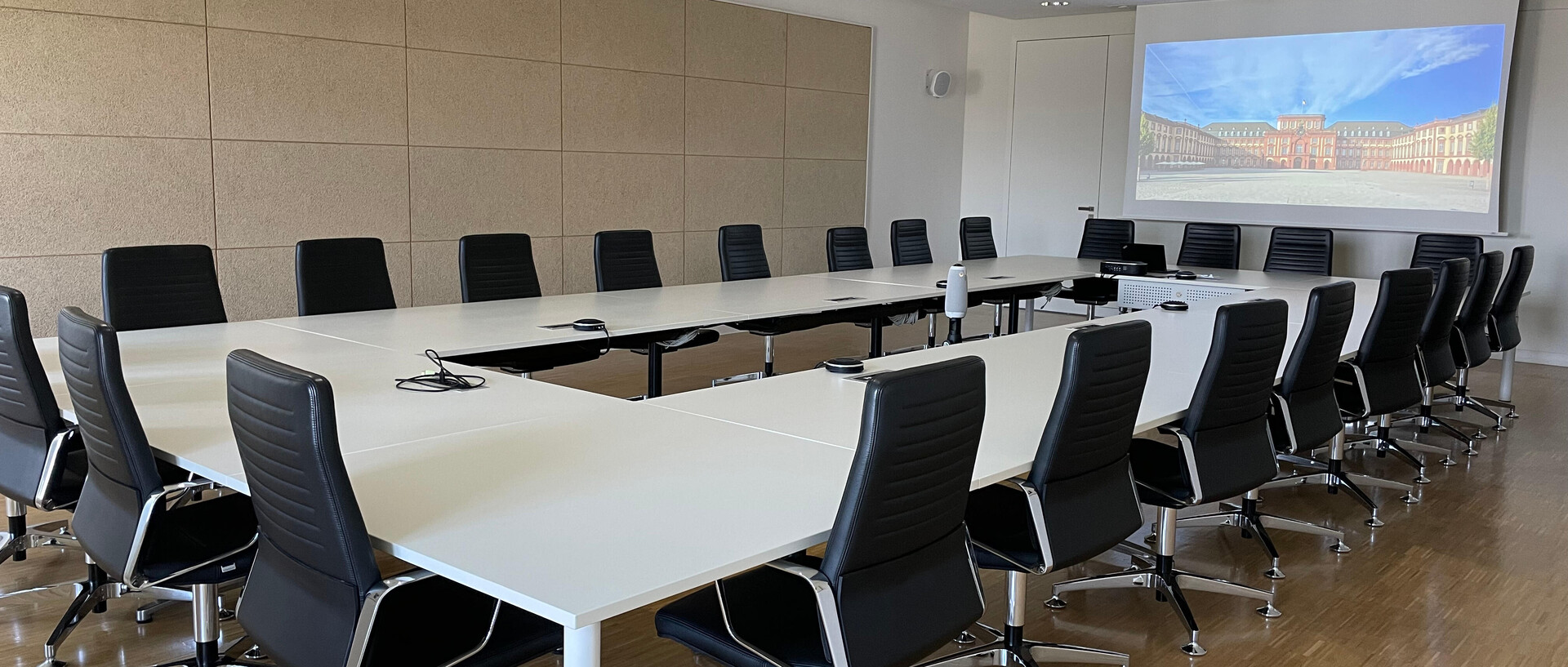 Moderner Besprechungsraum mit großem Tisch, an der Decke montiertem Beamer und Projektion auf eine Leinwand.