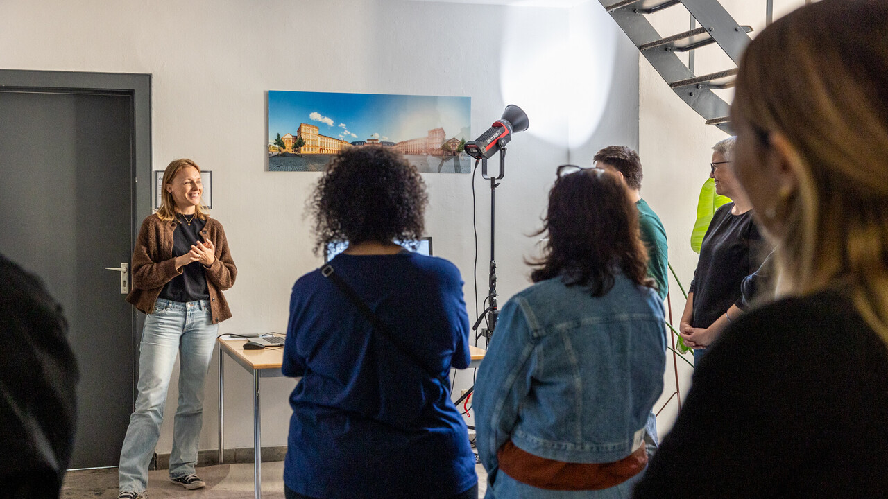 Eine Frau steht lächelnd im Foyer der Büros der Abteilung Marketing. Ihr gegenüber steht eine Gruppe Personen mit dem Rücken zur Kamera.