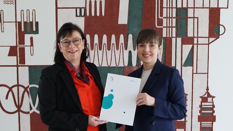 Brigitte Wecker and another woman are holding a certificate in front of a patterned wall.