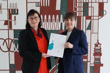 Brigitte Wecker and another woman are holding a certificate in front of a patterned wall.