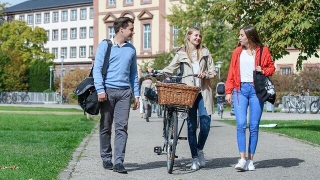 Drei Studierende laufen auf der Mensawiese. Die Person links hat kurze braune Haare und trägt ein blaues Oberteil. Die Person in der Mitte schiebt ihr Fahrrad neben sich her und hat lange blonde Haare. Sie trägt eine beige Jacke. Die Person rechts hat lange braune Haare und trägt ein weißes Oberteil mit einer roten Jacke.