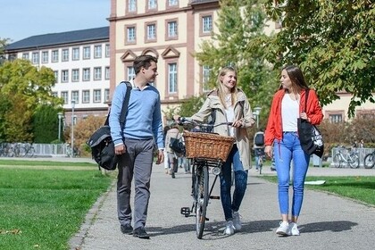 Drei Studierende laufen auf der Mensawiese. Die Person links hat kurze braune Haare und trägt ein blaues Oberteil. Die Person in der Mitte schiebt ihr Fahrrad neben sich her und hat lange blonde Haare. Sie trägt eine beige Jacke. Die Person rechts hat lange braune Haare und trägt ein weißes Oberteil mit einer roten Jacke.