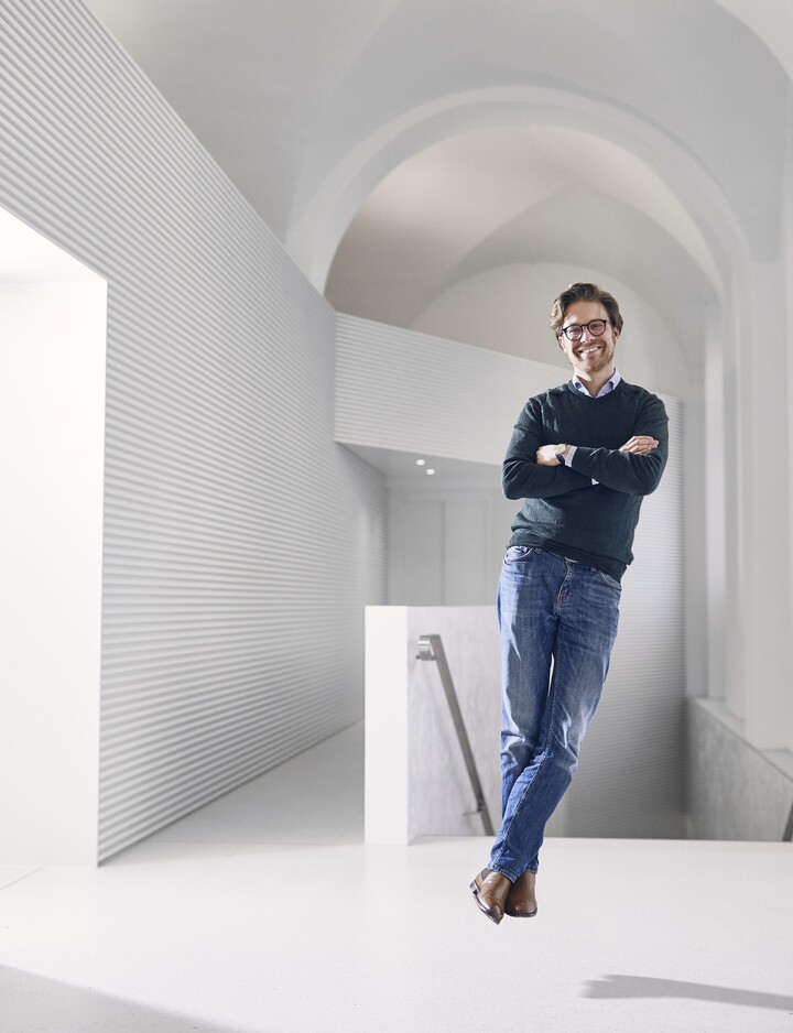 Mann mit Brille, Jeans, grauem Pullover und einem Hemd darunter steht schwebend über dem Boden in einem komplett weißen Raum und lächelt. 