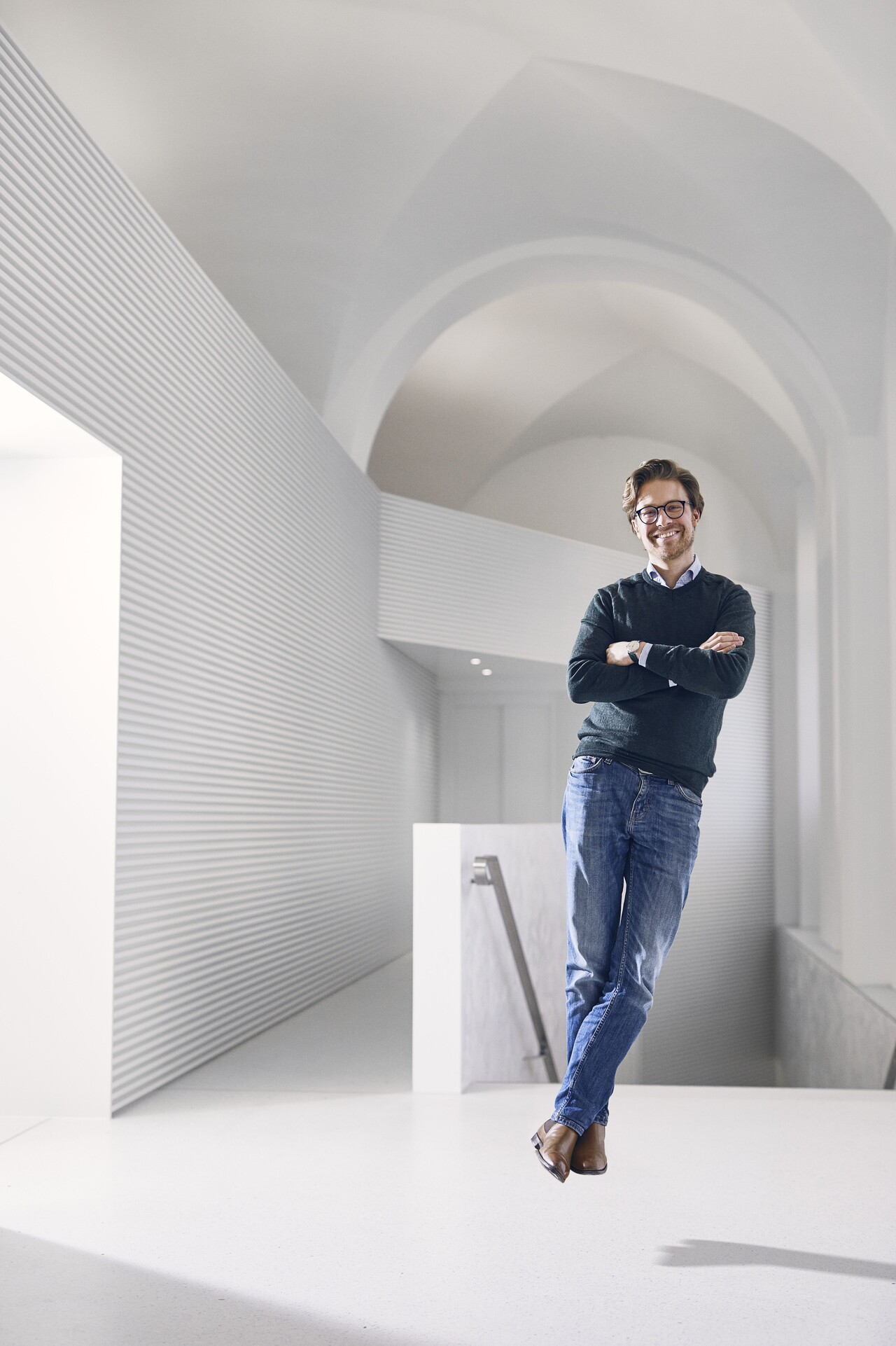 Mann mit Brille, Jeans, grauem Pullover und einem Hemd darunter steht schwebend über dem Boden in einem komplett weißen Raum und lächelt.