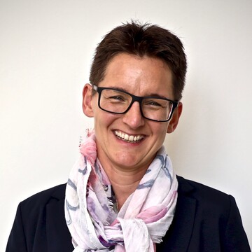 Andrea Kisel hat kurze braune Haare und eine dunkle Brille. Sie trägt einen hellrosanen Schal, ein weißes Oberteil und einen schwarzen Blazer.