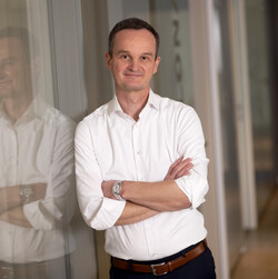 Rainer Wittwen hat kurze, dunkle Haare und trägt ein weißes Hemd mit hochgekrempelten Ärmeln. Er lehnt sich mit verschränkten Armen an eine Glaswand.