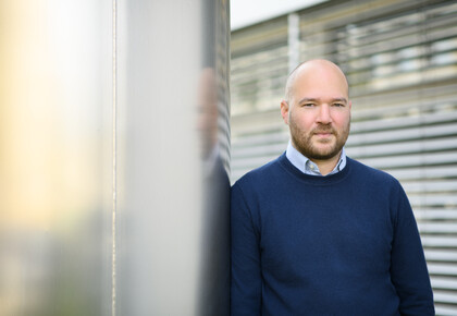 Eine Person trägt ein weiß-blau gestreiftes Hemd unter einem dunkelblauen Pullover und steht angelehnt an eine Säule. Die Person heißt Richard Traunmüller.