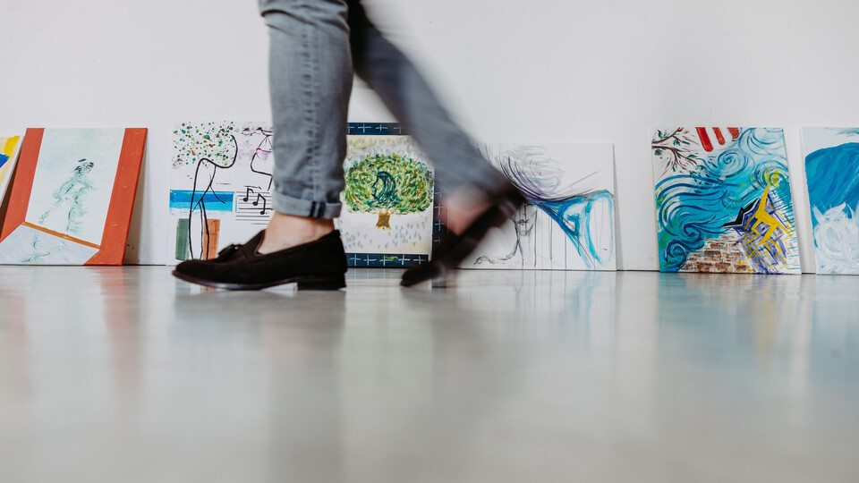 An eine Wand gelehnt stehen kleine, bunte Gemälde. Im Vordergrund sieht man zwei Beine in grauer Hose und mit schwarzen Ballerinas, die verwischt vorbeigehen.