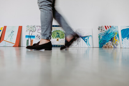 An eine Wand gelehnt stehen kleine, bunte Gemälde. Im Vordergrund sieht man zwei Beine in grauer Hose und mit schwarzen Ballerinas, die verwischt vorbeigehen.