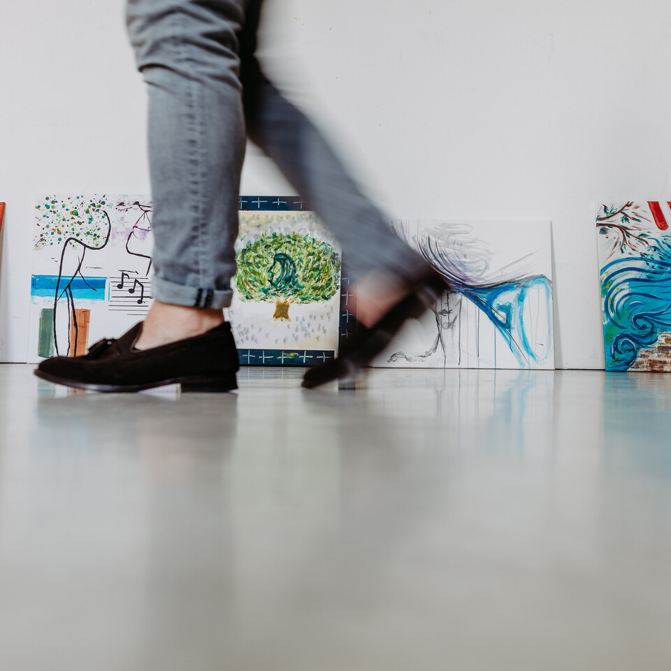 An eine Wand gelehnt stehen kleine, bunte Gemälde. Im Vordergrund sieht man zwei Beine in grauer Hose und mit schwarzen Ballerinas, die verwischt vorbeigehen.