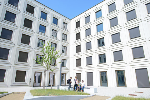 Innenhof des mit zahlreichen Fenstern gebauten Universitätsgebäudes B6 mit kleinen Grünflächen und im Hintergrund sitzen Studierende auf einer Bank.
