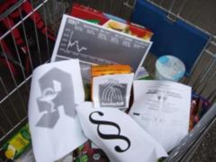 Ein Einkaufswagen mit verschiedenen Lebensmitteln und auf Papier gedruckte Symbole, darunter das Apothekenlogo, ein Paragraphenzeichen und ein Börsenauszug.