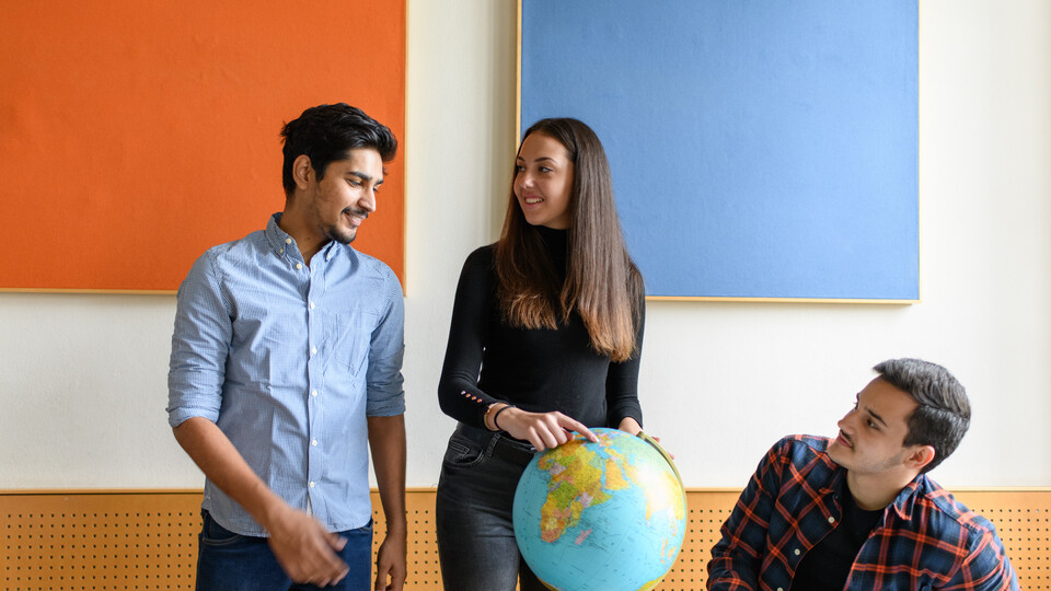 Drei Auslandsstudierende befinden sich mit einem Globus in einem Seminarraum. Eine Studentin zeigt auf ein Land auf dem Globus.