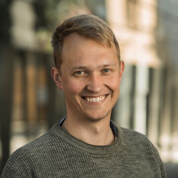 Leonard Wendering. Er trägt einen braunen Pullover und steht vor einem Gebäude mit hellen Steinwänden.