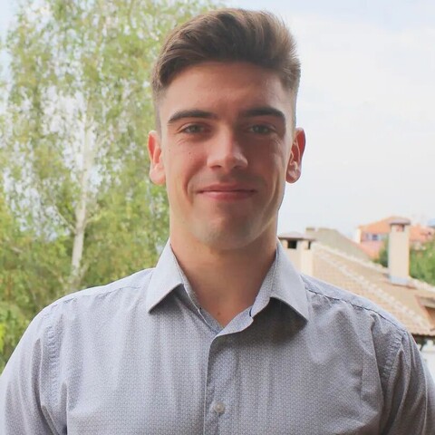 Georgi Paskalev has short brown hair and wears a gray shirt. You can see a few trees and houses in the background.
