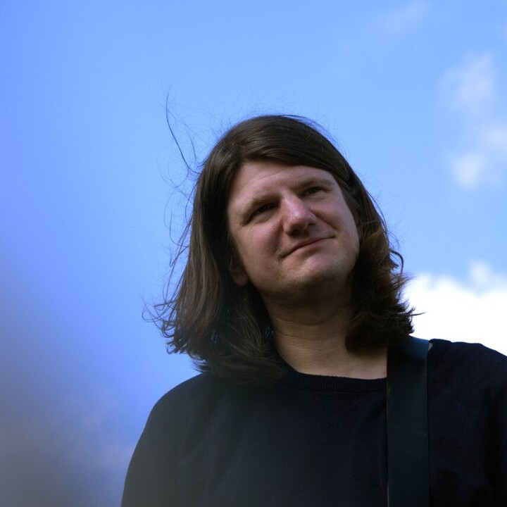 Ein Mann mit langen dunklen Haaren und einem schwarzen Outfit vor blauem Himmel.