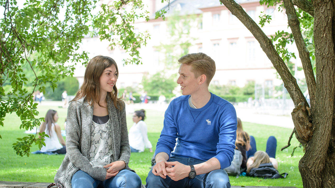 Eine Studentin und ein Student sitzen auf einer Mauer und lächeln sich an. Im Hintergrund sitzen einige Studierende auf der Mensawiese der Universität Mannheim.