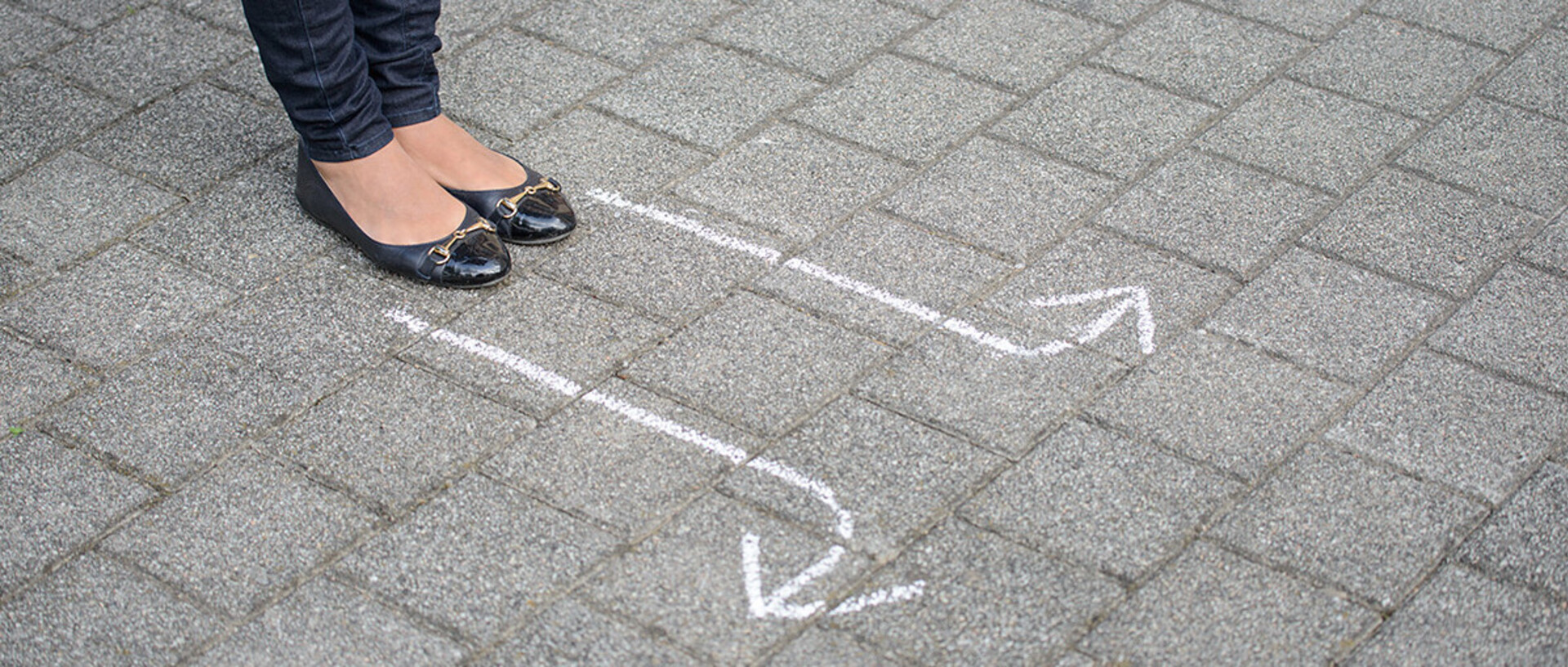 Beine und Füße einer Frau auf dem Boden stehend. Vor ihr sind mit Kreide Pfeile nach links und rechts auf den Boden gemalt.