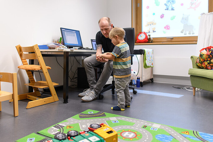 Father and son are sitting in a playroom, the father is showing something to his son.