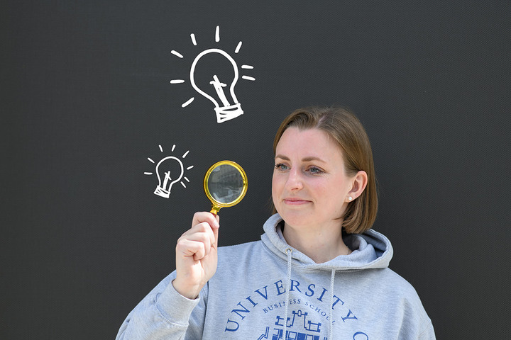 A young woman is looking for information about the degree process. She is holding a lupe and next to her are light bulbs.