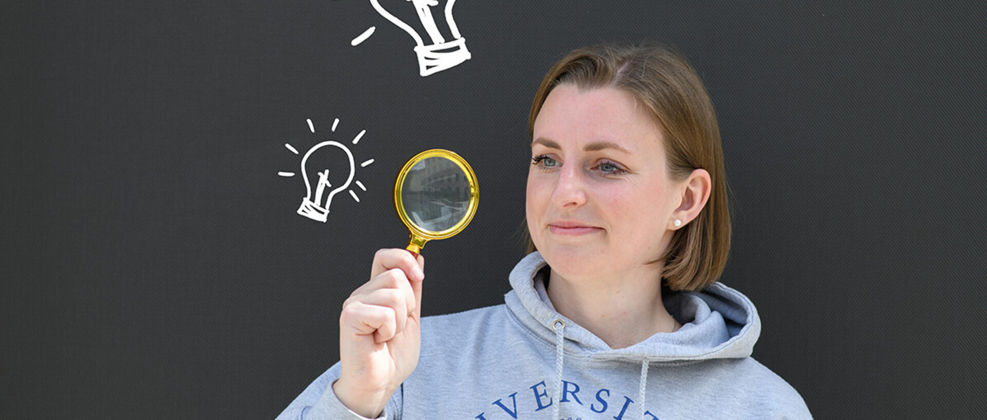 A young woman is looking for information about the degree process. She is holding a lupe and next to her are light bulbs.