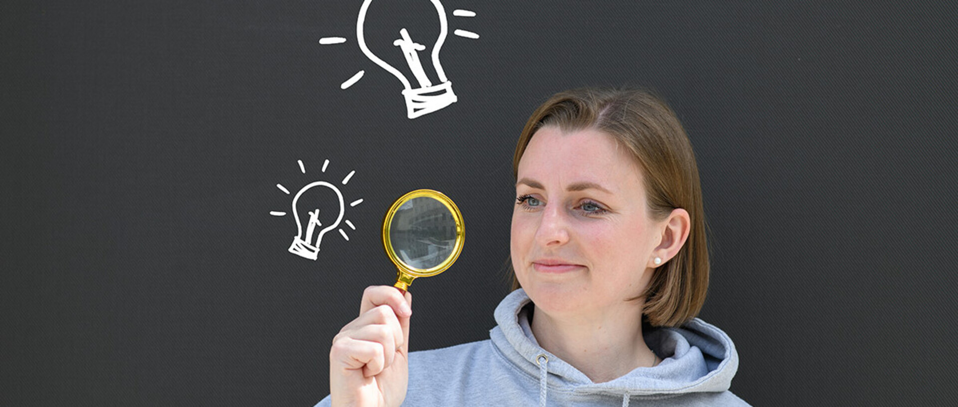 A young woman is looking for information about the degree process. She is holding a lupe and next to her are light bulbs.