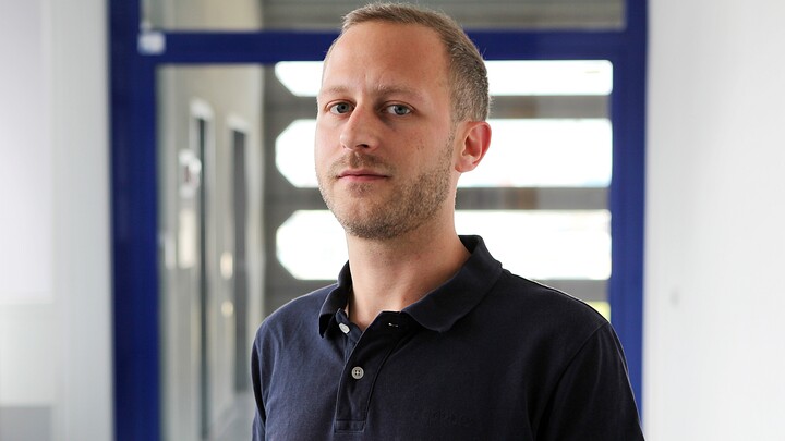 Eine Person trägt einen dunkelblauen Pullover und steht vor einer Glastüre. Die Person heißt Christian Glässel.