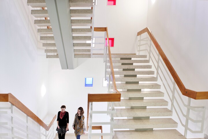 Zwei Frauen laufen in einem hell erleuchteten Treppenhaus die Treppen herauf. Der Handlauf ist aus Holz, das Geländer aus weißem Metall und die Treppenstufen aus hellem Stein.