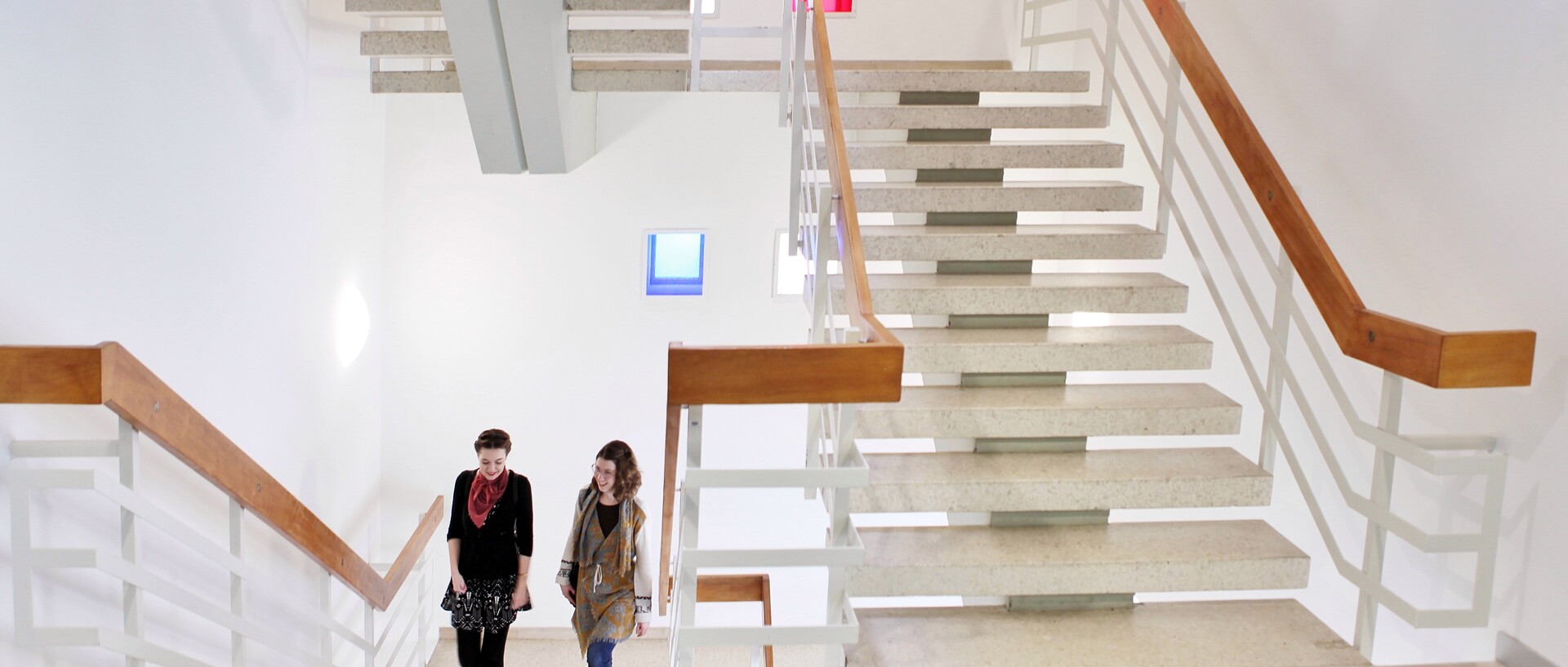 Zwei Frauen laufen in einem hell erleuchteten Treppenhaus die Treppen herauf. Der Handlauf ist aus Holz, das Geländer aus weißem Metall und die Treppenstufen aus hellem Stein.