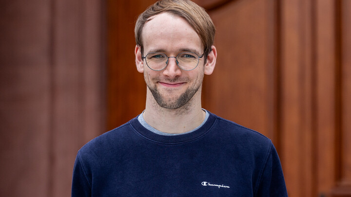Portrait of Max Riegel. He is wearing a dark blue sweater and is standing in front of a door of Mannheim Castle.