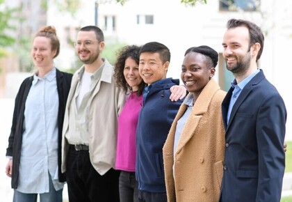 Six people are standing in a row. They have different genders and cultural backgrounds.