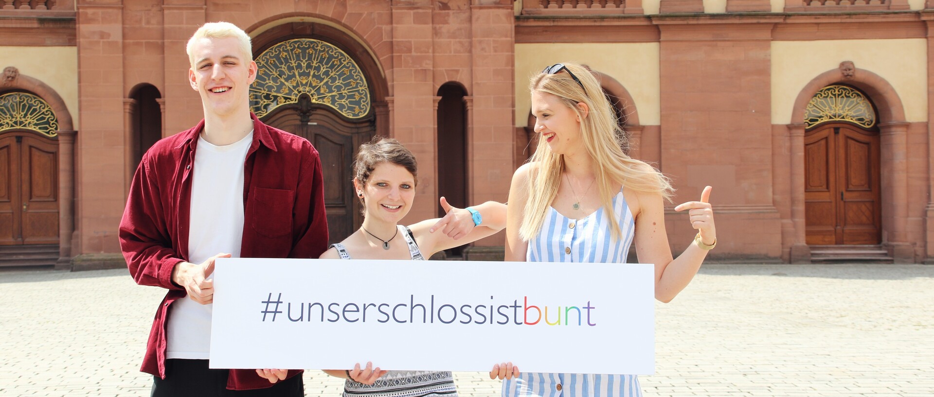 Drei Personen stehen auf dem Ehrenhof und halten ein Schild mit der Aufschrift "#unser schloss ist bunt" in die Höhe.