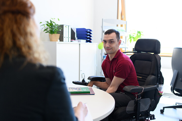 Eine Mitarbeiterin berät einen Studenten, der in einem Elektrorollstuhl sitzt.